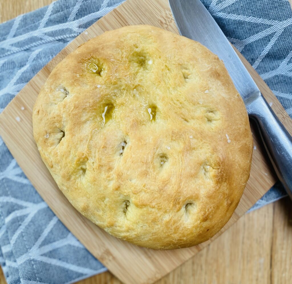 A baked focaccia bread, on a wooden board, with a stainless steel knife on the board. A grey, patterned tea-towel underneath. 