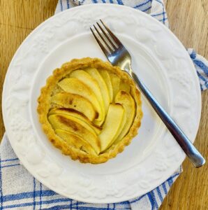 an individual French apple tart on a white plate on top of a blue patterned tea-towel, with a stainless steel fork