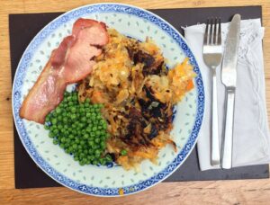 potato and onion hash, with bacon, peas and ketchup, on a blue patterned plate, with a white napkin and stainless cutlery