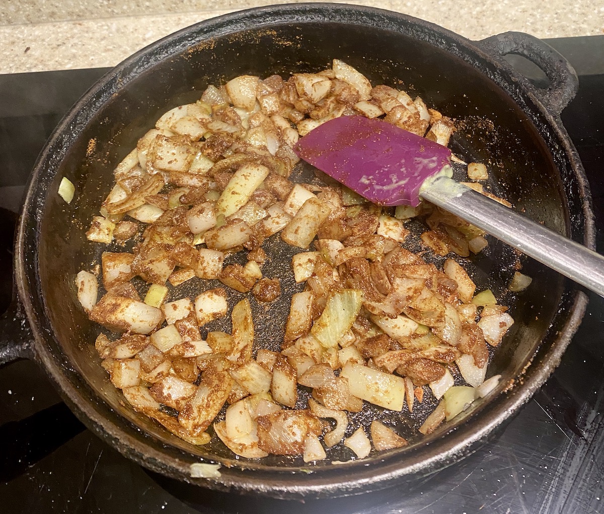 a cast iron frying pan with fried onions and spices. A purple silicone spatula in top right.