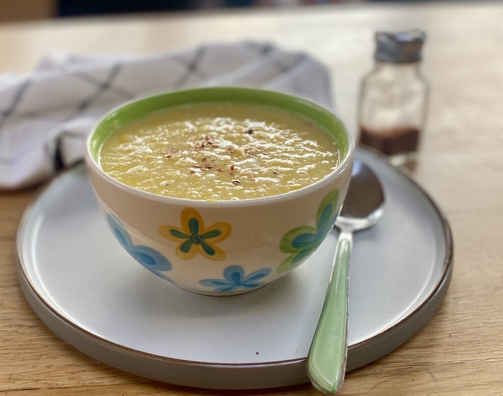 sweetcorn soup in a multi coloured bowl, standing on a neutral coloured plate with a pale green handled spoon. A grey checked tea towel is nearby. 
