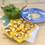 baked puff pastry, red pesto, butternut squash and chicken tart. 2 plates and cutlery stacked nearby. Glass bowl of salad leaves on the top left.