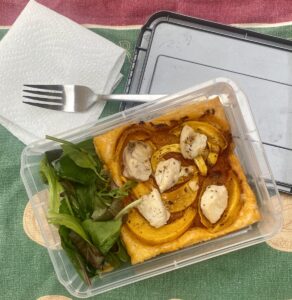 a savory pumpkin tart in a transparent lunch box, with green leaves. A lid and fork are nearby, on a paper napkin, all resting on a green tea towel.