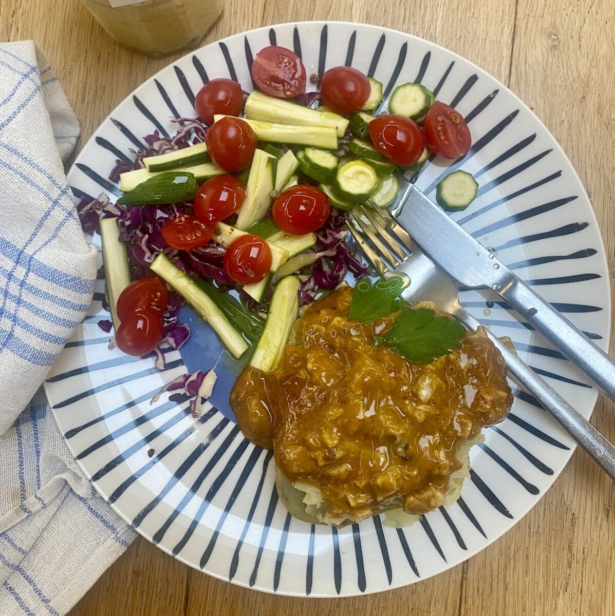 Coronation chicken on a baked potato, with salad, on a blue striped plate.