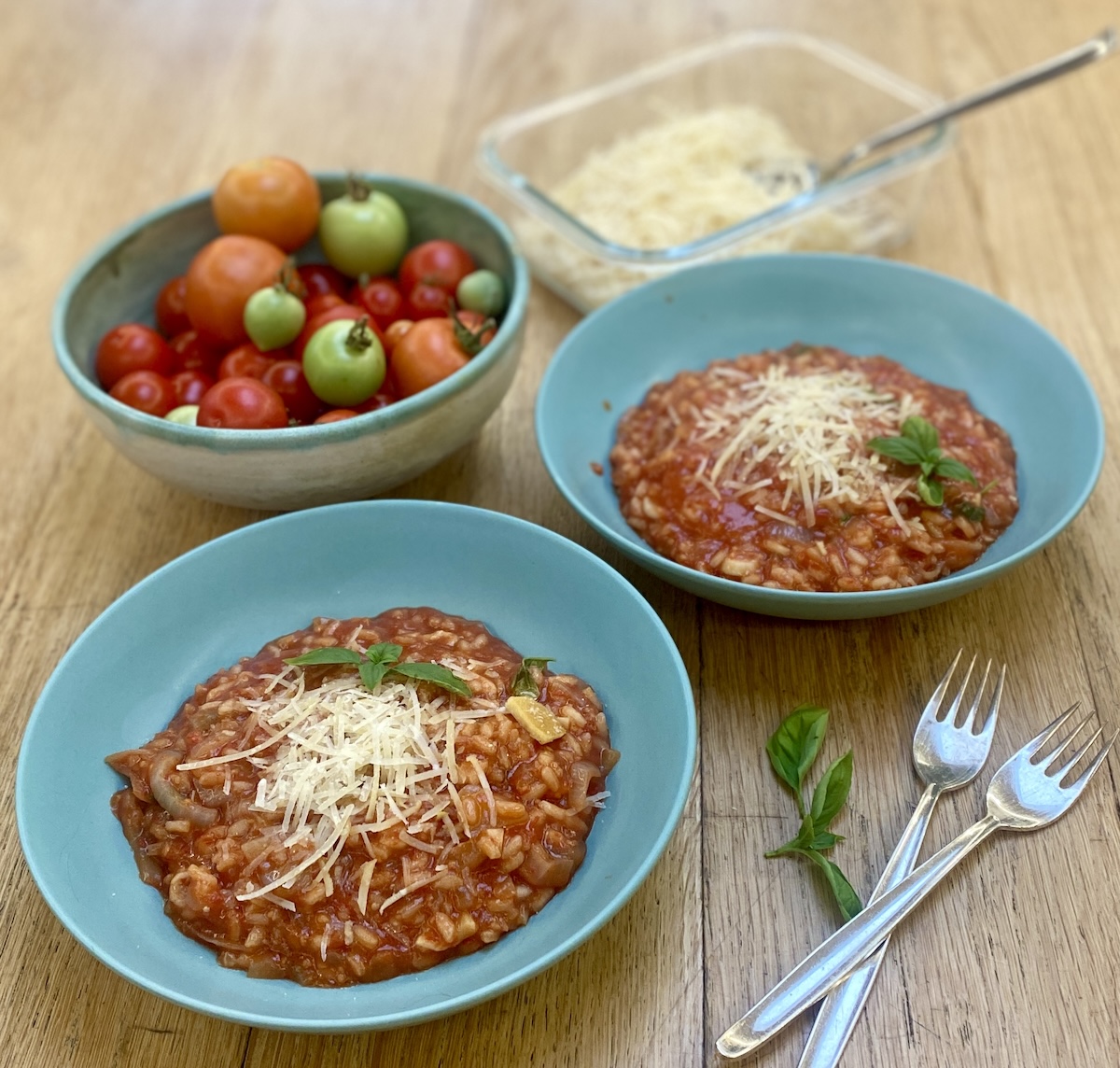 two portions of tomato risotto in duck egg blue bowls, with a dish of fresh tomatoes, forks and a dish of parmesan. 