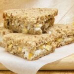 a chicken and sweetcorn sandwich, cut into quarters, on greaseproof paper and a wooden board.