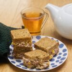 a tuna and sweetcorn sandwich, cut inti 4 squares, on a blue patterned tea-plate with a tea-pot and cup in the background.