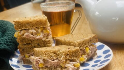 a tuna and sweetcorn sandwich, cut inti 4 squares, on a blue patterned tea-plate with a tea-pot and cup in the background.