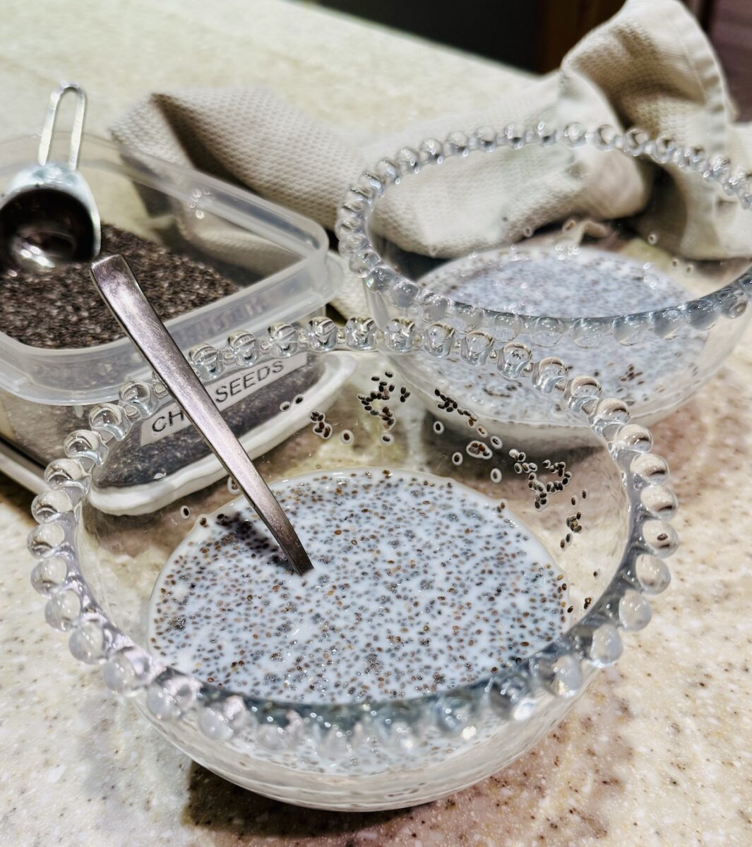 chia seeds soaking in two glass bowls, with bobbles round the top. One bowl has a tea spoon in it, there is a plastic storage container containing chia seeds. a tea -towel is folded next to one of the bowls. 