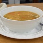 Frozen mixed vegetable soup in a white bowl, on a white plate with a spoon, and a folded tea-towel on the left