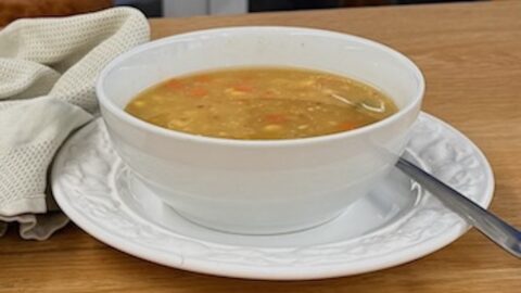 Frozen mixed vegetable soup in a white bowl, on a white plate with a spoon, and a folded tea-towel on the left