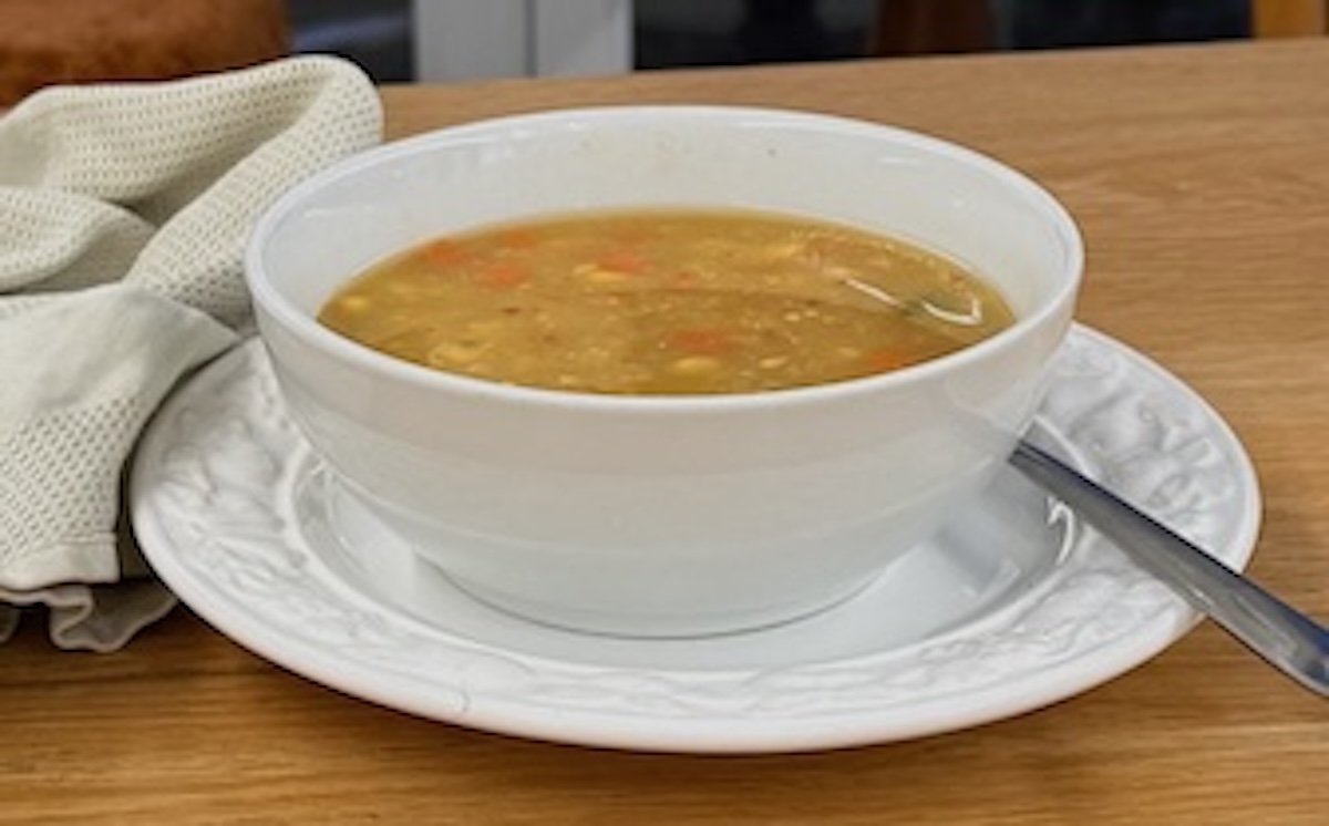 Frozen mixed vegetable soup in a white bowl, on a white plate with a spoon, and a folded tea-towel on the left